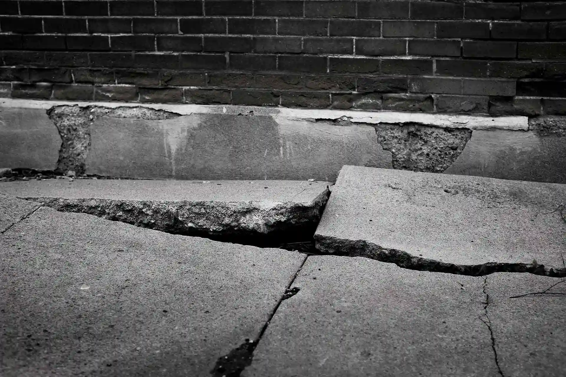 A broken and damaged sidewalk against a brick wall