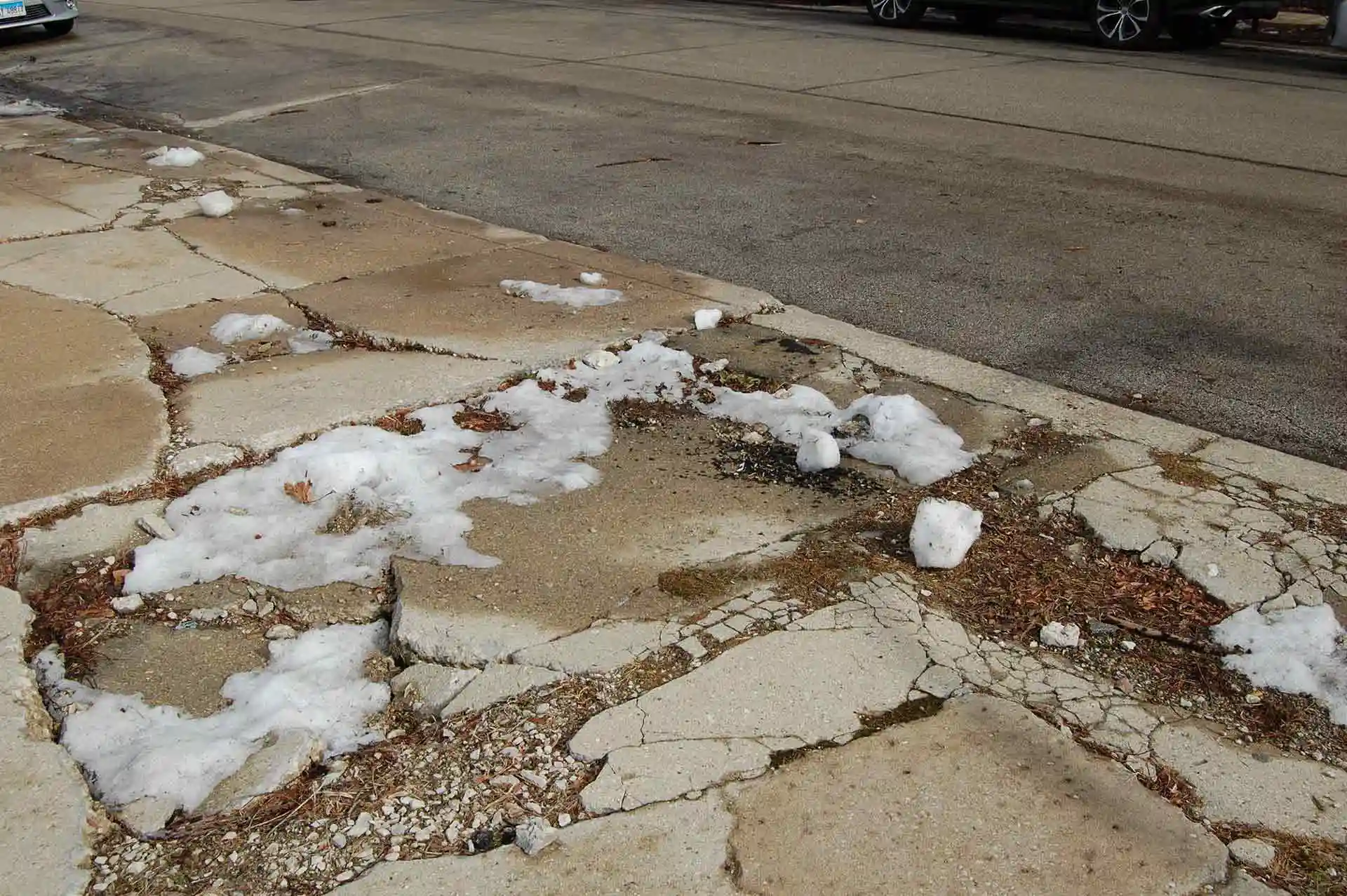 A broken and damaged sidewalk with patches of snow on it